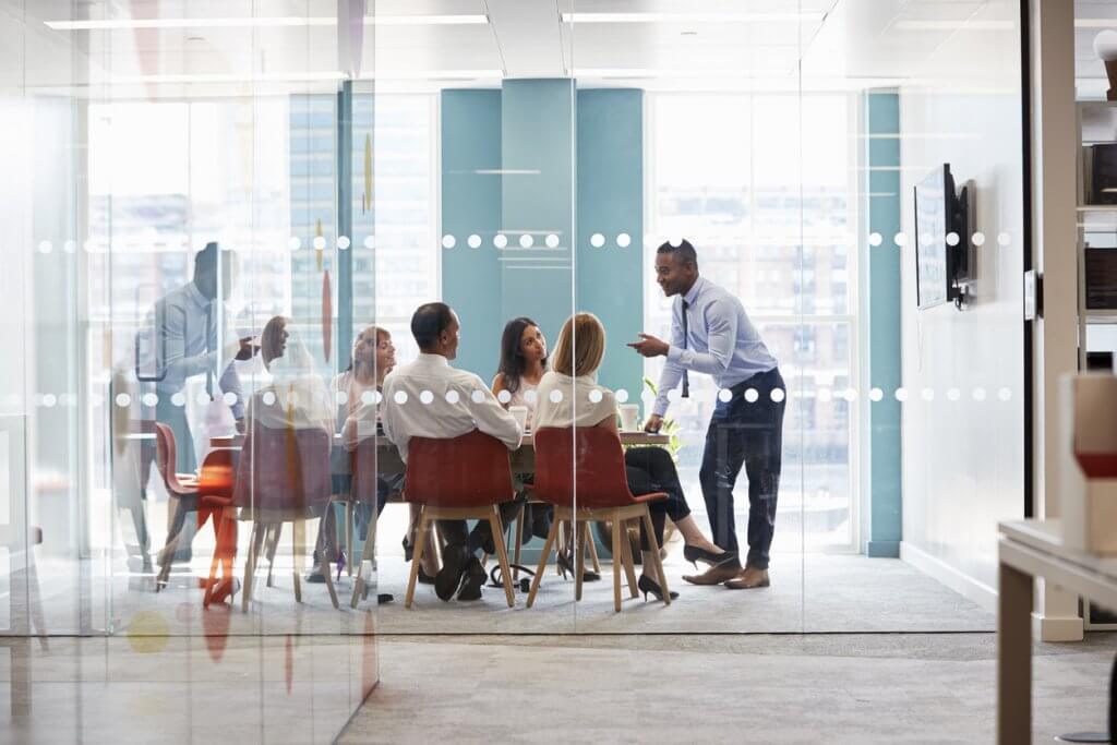 man-speaking-to-team-of-employees-seated-in-red-chairs-at-conference-table-in-room-with-glass-wall